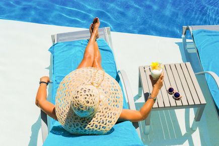 Frau mit großem Strohhut liegt auf blauem Liegestuhl neben Pool, hält Cocktail mit Blume, neben ihr Tisch mit Sonnenbrille