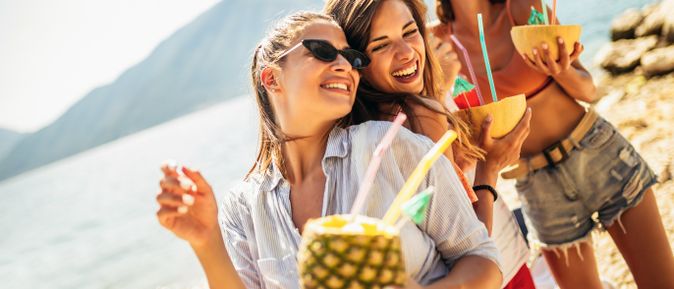 Junge Frauen lachen und tanzen am Strand, halten bunte Cocktails in Früchten wie Ananas und Melone in den Händen
