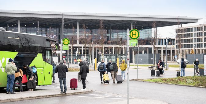 Mehrere Personen mit Gepäck stehen und gehen an einer Bushaltestelle vor dem Terminal des Flughafens Berlin-Brandenburg.