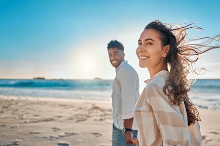 Lächelndes Paar spaziert bei Sonnenuntergang am Strand entlang, mit Wind im Haar und Meereskulisse