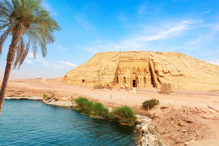 Abu-Simbel-Tempel mit vier großen sitzenden Pharaonenstatuen am Fels, davor Wasser und Palmen
