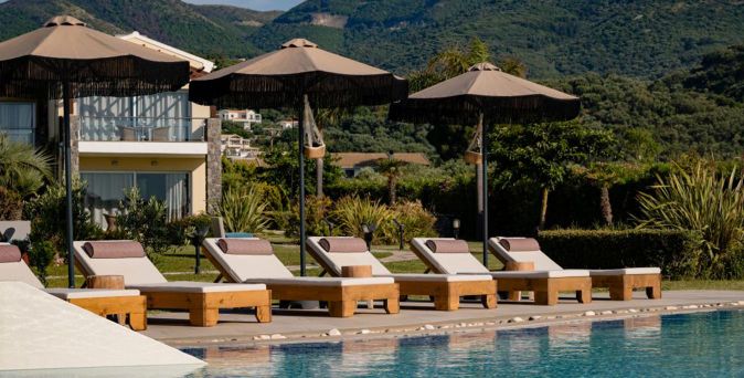 Reihe von Liegestühlen mit Polstern und Sonnenschirmen am Pool vor einem Hotelgebäude und bewaldeten Hügeln