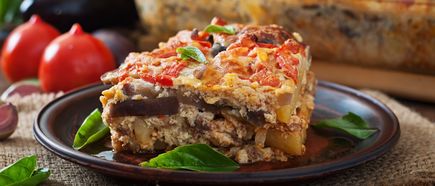 Nahaufnahme eines Stücks Moussaka mit sichtbaren Auberginen- und Tomatenschichten auf einem Teller mit grünen Blättern im Hintergrund.