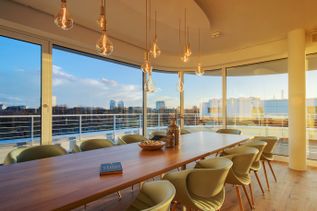 Skylounge in Gebäude 3 von schauinsland-reisen mit großem Holztisch, mehreren Sitzplätzen, modernen Pendelleuchten und Panoramablick über die umliegende Stadt durch bodentiefe Fenster
