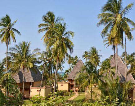 Mehrere strohgedeckte Bungalows umgeben von Palmen und üppiger Vegetation unter blauem Himmel im Neptune Pwani Beach Resort & Spa auf Sansibar.