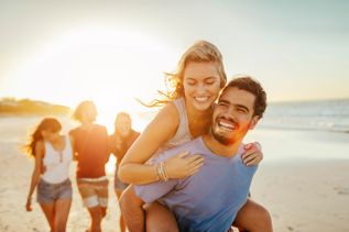 Frau lässt sich lachend von Mann am Strand tragen, während weitere Freundinnen und Freunde hinter ihnen durch den warm beleuchteten Sand laufen