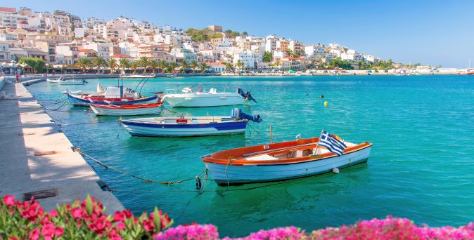 Mehrere kleine Boote mit griechischer Flagge liegen an einer Hafenmauer, im Hintergrund Häuser an einem Hang und blühende pinke Blumen im Vordergrund.