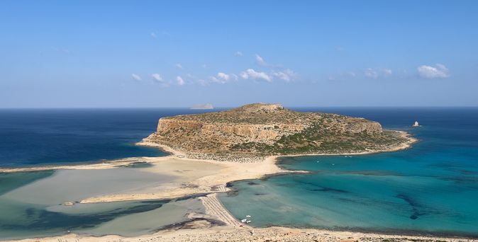 Eine sandige Landzunge verbindet den Strand mit einer flachen Felseninsel, umgeben von türkisfarbenen und dunkleren Meeresbereichen. Der Himmel ist klar und blau, und die Landschaft wirkt weit und unbebaut.