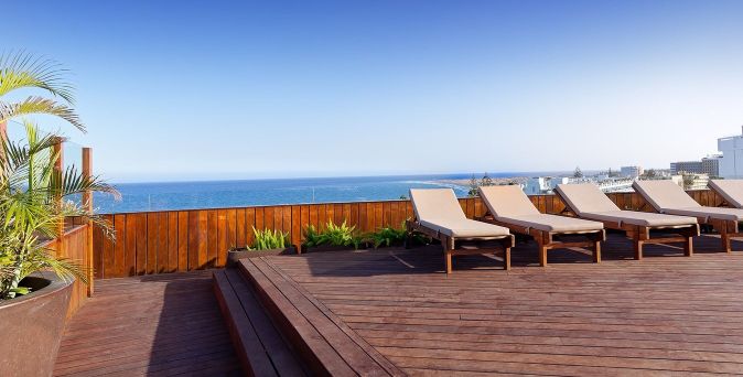 Holzterrasse mit fünf Liegen, Blick auf das Meer und die Küste, Palmen und Pflanzen im Vordergrund.