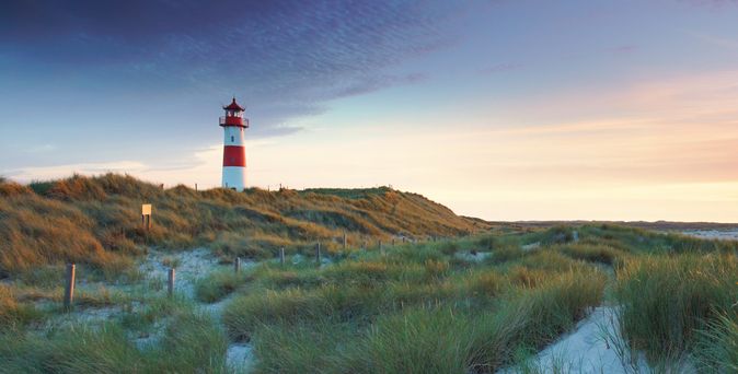 Leuchtturm auf einer Dünenlandschaft mit Gras unter einem bewölkten Himmel bei Sonnenuntergang