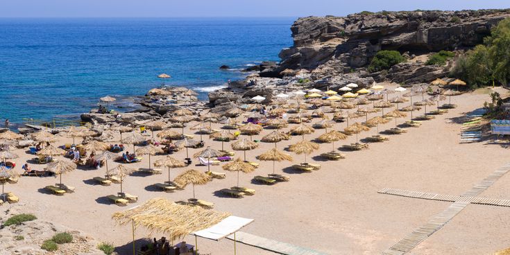Strand mit Reihen von Sonnenschirmen aus Stroh und gelben Sonnenschirmen, felsige Küste und blaues Meer im Hintergrund.