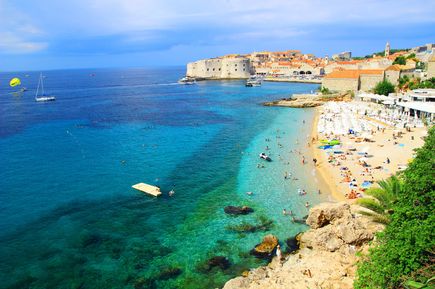 Blick über den Banje-Strand mit Badegästen und türkisblauem Meer auf die historische Altstadt von Dubrovnik mit ihren mächtigen Stadtmauern