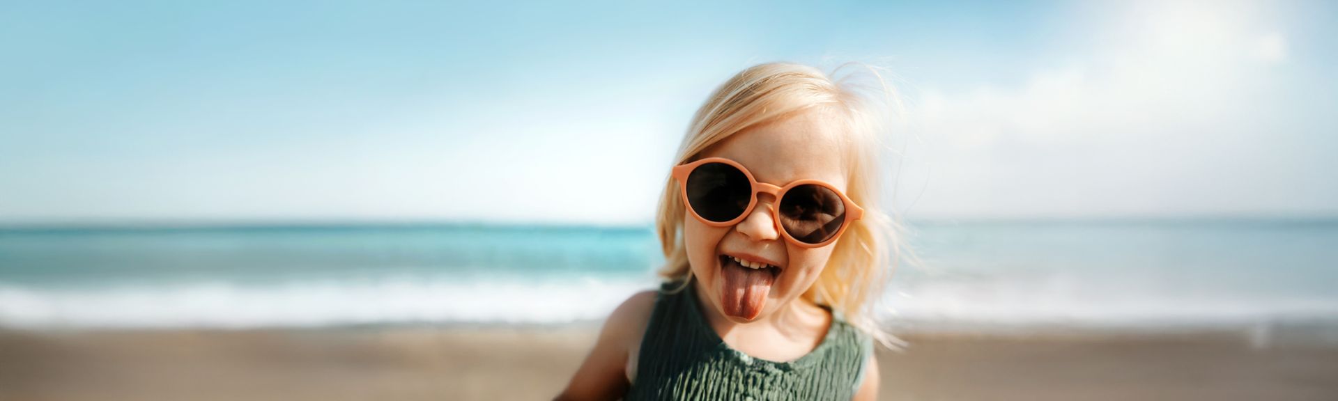 Ein kleines Kind mit großer, orangefarbener Sonnenbrille steht am Strand vor dem Meer, bei hellem Sonnenlicht und leichtem Wind und streckt dabei die Zunge heraus, während der Sandstrand und die Meereswellen im Hintergrund sichtbar sind.