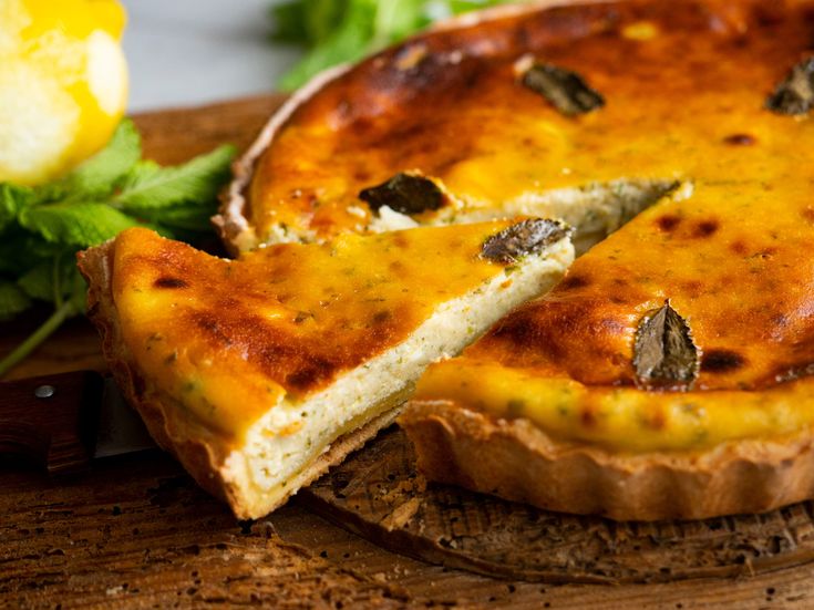 Angeschnittener Flaó-Kuchen mit goldbrauner Oberfläche auf einem Holzbrett, sichtbare cremige Füllung, dekoriert mit Kräutern, Zitronen und Grün im Hintergrund
