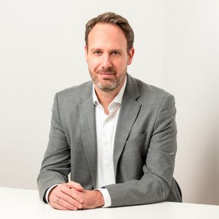 orträt von Sascha Leutner, sitzend an einem Tisch vor hellem Hintergrund, gekleidet in ein graues Sakko und weißes Hemd, mit direktem Blick in die Kamera