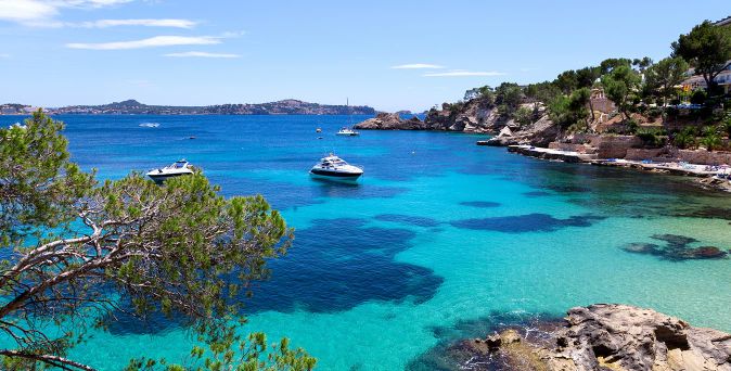 Bucht mit klarem türkisfarbenem Wasser, zwei Booten und bewaldeten Felsen im Hintergrund