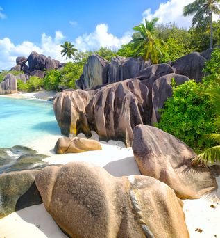 Strand mit großen Granitfelsen, weißem Sand, türkisfarbenem Wasser und Palmen unter blauem Himmel auf den Seychellen