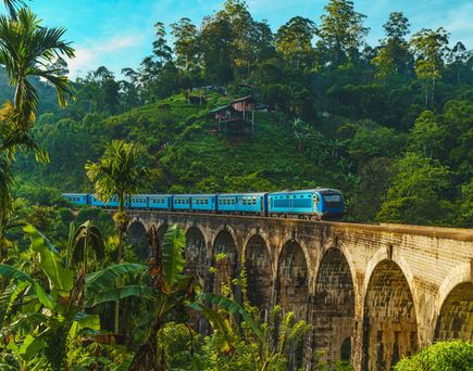 Ein langer blauer Zug fährt über die historische Neun-Bogen-Steinbrücke in Ella auf Sri Lanka, umgeben von dichtem grünen Dschungel.