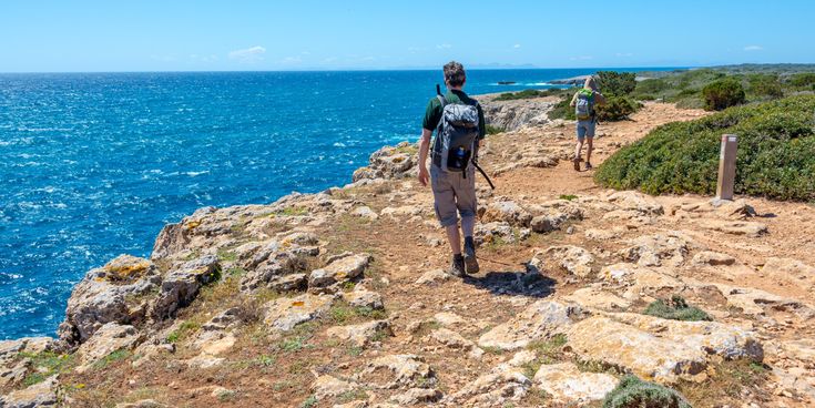 Zwei Wanderer mit Rucksäcken gehen auf einem felsigen Küstenpfad mit Blick auf das blaue Meer