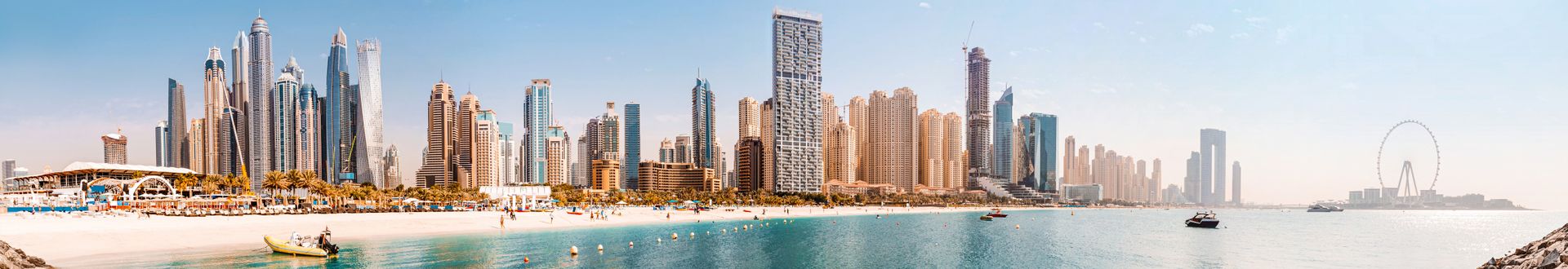 Panoramaaufnahme der Skyline von Dubai mit modernen Wolkenkratzern, einem Strand und dem Dubai Eye im Hintergrund. Klare Himmel und ruhiges Wasser.