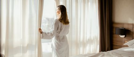 Person im weißen Bademantel steht am Fenster eines Hotelzimmers und blickt durch lichtdurchflutete Vorhänge auf die Umgebung