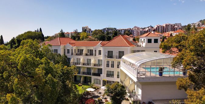 Hotelgebäude mit roten Ziegeldächern und einem überdachten Pool auf einer Terrasse, umgeben von Bäumen und weiteren Gebäuden im Hintergrund