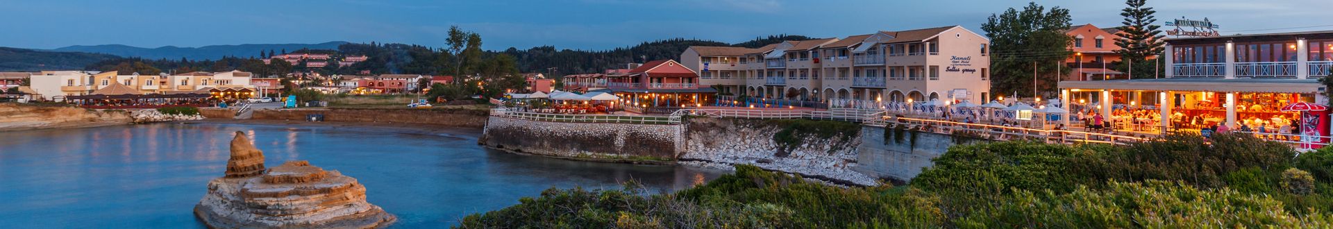 Panoramablick auf eine Küstenstadt mit Felsen im Wasser, mehreren Gebäuden und beleuchteten Restaurants am Abend.