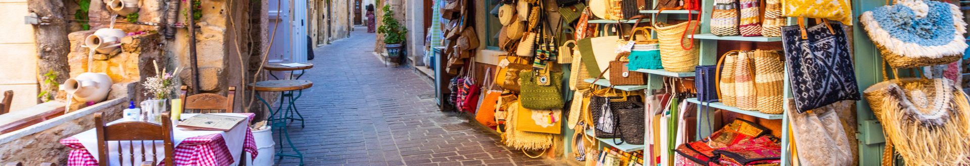 Eng gepflasterte Einkaufsstraße mit Tischen, Stühlen und bunten Warenständen an den Seiten, mediterranes Flair.