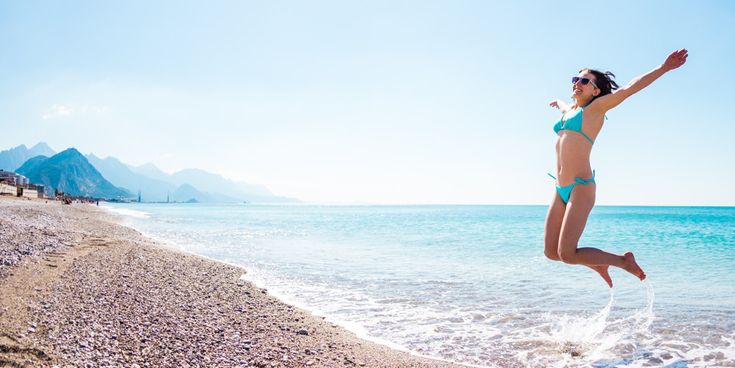 Frau springt lachend am Strand, mit klarem Meer und Bergkulisse im Hintergrund