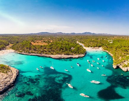 Blick auf die Bucht Cala Mondragó auf Mallorca mit türkisfarbenem Wasser, bewaldeten Küsten, Sandstrand und zahlreichen Booten in einer naturbelassenen Umgebung