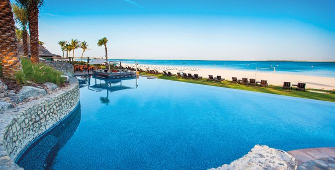 nfinity-Pool des JA Palm Tree Court in Dubai mit Blick auf das Meer, umgeben von Palmen, Sonnenliegen und direktem Zugang zum Sandstrand bei klarem Himmel