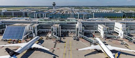 Zwei Verkehrsflugzeuge an den Gates von Terminal 2 am Flughafen München, Vorfeld mit Abfertigungsfahrzeugen, Fluggastbrücken und Terminalgebäude mit Solarpanels sowie Tower im Hintergrund