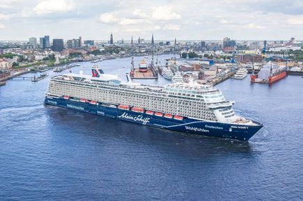 Das Kreuzfahrtschiff „Mein Schiff 7“ fährt durch den Hamburger Hafen, umgeben von Werftanlagen, Kranschiffen und der städtischen Skyline im Hintergrund.