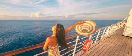 Frau steht auf dem Deck eines Kreuzfahrtschiffs, hält einen Hut in die Luft und blickt auf das weite Meer bei Sonnenlicht