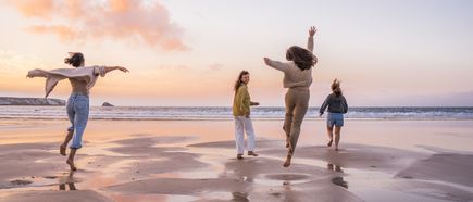 Vier Personen laufen und springen am Strand entlang, während sich farbiges Abendlicht im nassen Sand spiegelt und Wellen im Hintergrund an die Küste rollen
