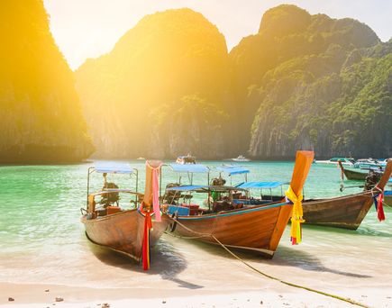 Drei traditionelle thailändische Langheckboote im türkisfarbenen Wasser vor einer Sandküste und bewaldeten Kalksteinfelsen bei Sonnenuntergang