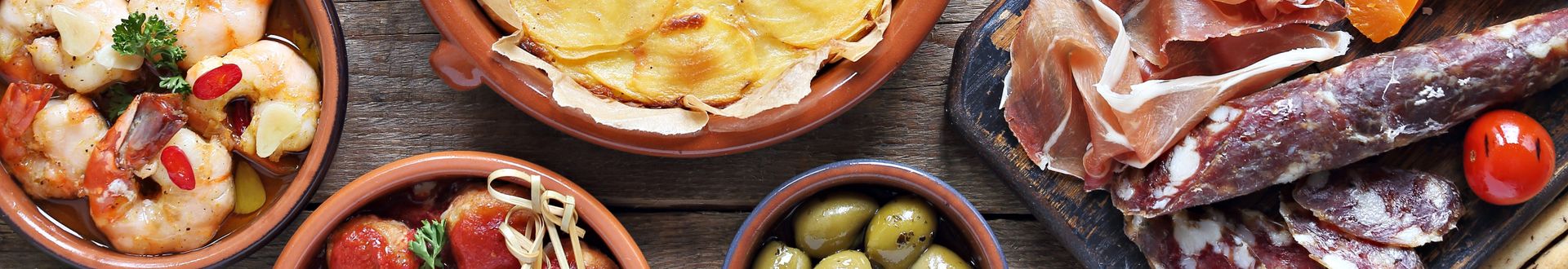 Spanische Tapas-Auswahl auf Holztisch: Garnelen, Auflauf mit Kartoffelscheiben, grüne und schwarze Oliven, gefüllte Paprika, luftgetrockneter Schinken und Wurst, Pistazien