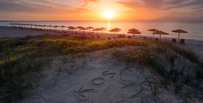 Strand mit Sanddünen, auf denen das Wort 'KOS' geschrieben ist, davor eine Reihe von Sonnenschirmen und der Sonnenuntergang über dem Meer