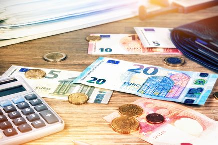 Mehrere Euro-Banknoten in den Stückelungen 5, 10, 20 und 50 liegen verstreut auf einem Holztisch neben Münzen und einem Taschenrechner.