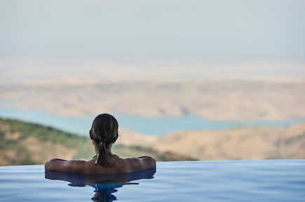 Frau lehnt entspannt am Rand eines Infinity-Pools und schaut über eine weite Berglandschaft mit Seen