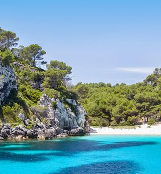 Klares türkisfarbenes Wasser in einer Bucht umgeben von steilen Felsen und grünen Kiefern auf Menorca