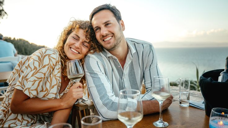 Zwei Personen sitzen an einem Tisch in einem Restaurant mit Meerblick und Weingläsern vor sich, während die Küstenlandschaft im Hintergrund zu sehen ist