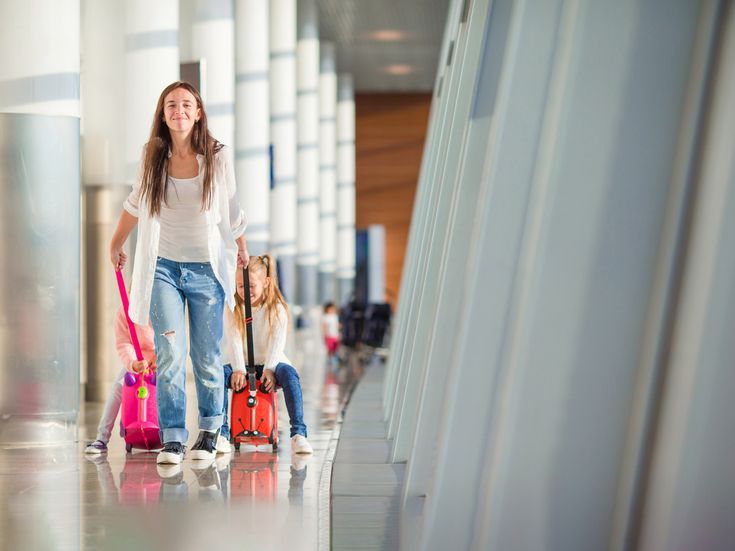 Frau mit zwei Kindern und Koffern läuft lächelnd durch die helle Abflughalle eines Flughafens