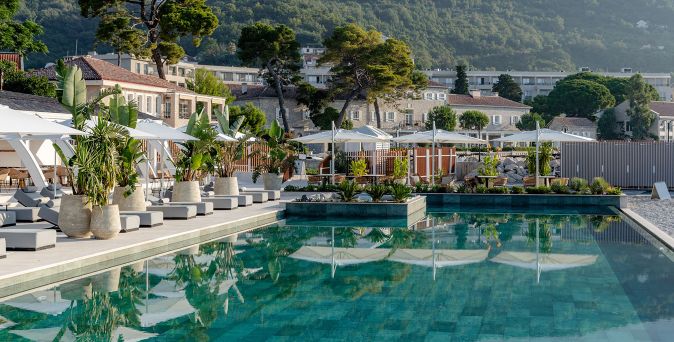 Großer Außenpool mit Liegen und Sonnenschirmen am Lazure Hotel & Marina Montenegro, umgeben von mediterraner Vegetation und Gebäuden vor bewaldeten Hügeln