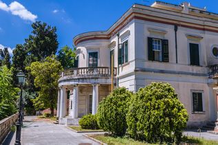 Neoklassizistisches Gebäude mit Säulen und Balkon umgeben von Bäumen und Sträuchern unter blauem Himmel