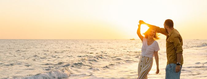 Mann und Frau tanzen lachend im seichten Wasser am Strand bei Sonnenuntergang