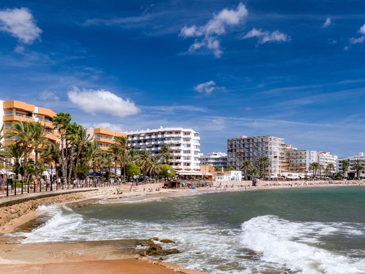 Strandpromenade von Santa Eulalia auf Ibiza mit Palmen, Hotels und Restaurants entlang der Küste sowie Wellen, die ans Ufer schlagen