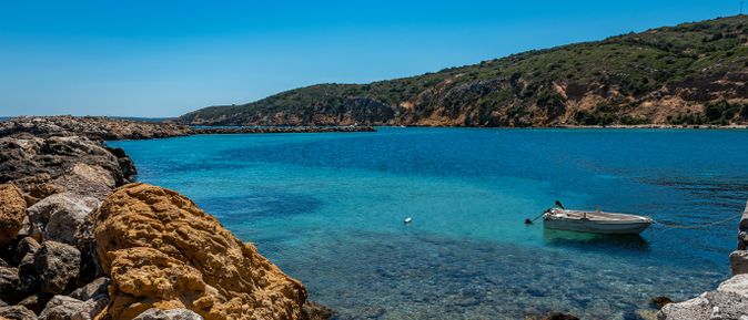 Klares türkisblaues Wasser an felsiger Küste mit grün bewachsenen Hügeln im Hintergrund und einem kleinen weißen Boot auf dem Wasser.