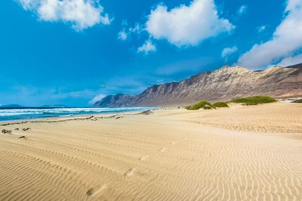 Playa de Famara auf Lanzarote mit weitläufigem Sandstrand, Dünen im Vordergrund, Atlantikbrandung und dem steil aufragenden Famara-Massiv unter blauem Himmel