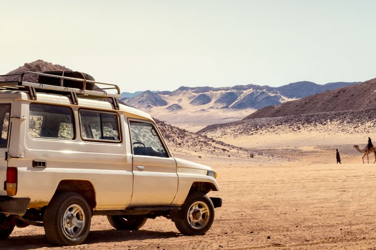 Ein geländegängiger Jeep steht in einer sandigen Wüstenlandschaft, im Hintergrund Berge und ein Reiter auf einem Kamel